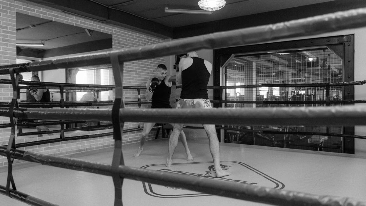 Black and white image of two men sparring in a boxing ring inside a gym.